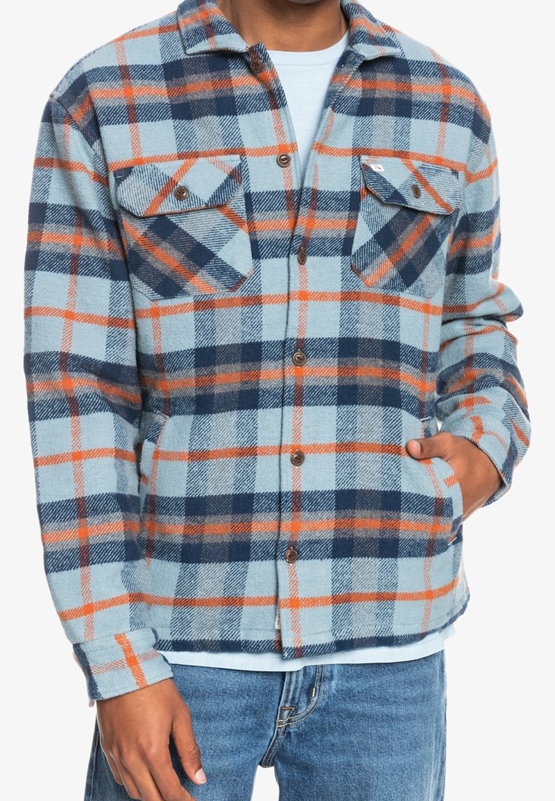 Man wearing light blue, navy, and orange plaid shirt jacket, with hand in side pocket, paired with blue jeans and a light blue undershirt.