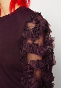 Ripresa ravvicinata di un top viola con maniche trasparenti adornato da applicazioni di fiori in tessuto, indossato da una persona con capelli rossi.