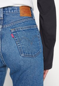 Jeans en denim bleu foncé avec un design classique, présentant un motif de couture distinctif sur la poche arrière et une étiquette en cuir à la taille.