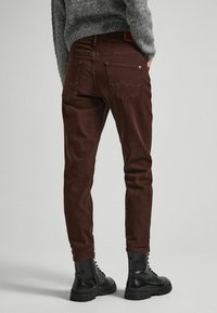 Jeans marroni aderenti con tasche posteriori e dettagli di cuciture sottili, abbinati a stivali neri con lacci. Maglione grigio testurizzato visibile sopra.