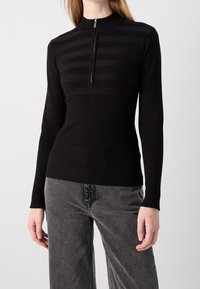 Schwarzer Strickoberteil mit hohem Kragen und Reißverschluss; verfügt über horizontale Streifen im oberen Bereich und eine gerippte Textur an Ärmeln und Körper.