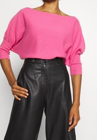 Rosa übergroßer Pullover mit weitem Halsausschnitt und Dreiviertelärmeln, kombiniert mit hoch geschnittenen schwarzen Lederhosen. Glatte Textur.