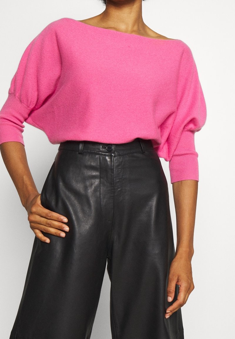 Rosa übergroßer Pullover mit weitem Halsausschnitt und Dreiviertelärmeln, kombiniert mit hoch geschnittenen schwarzen Lederhosen. Glatte Textur.