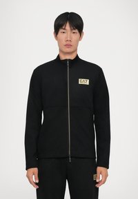 Uomo che indossa una giacca nera con zip e pantaloni con toppe del logo EA7 Emporio Armani sul petto e sulla coscia, in piedi davanti a uno sfondo semplice.