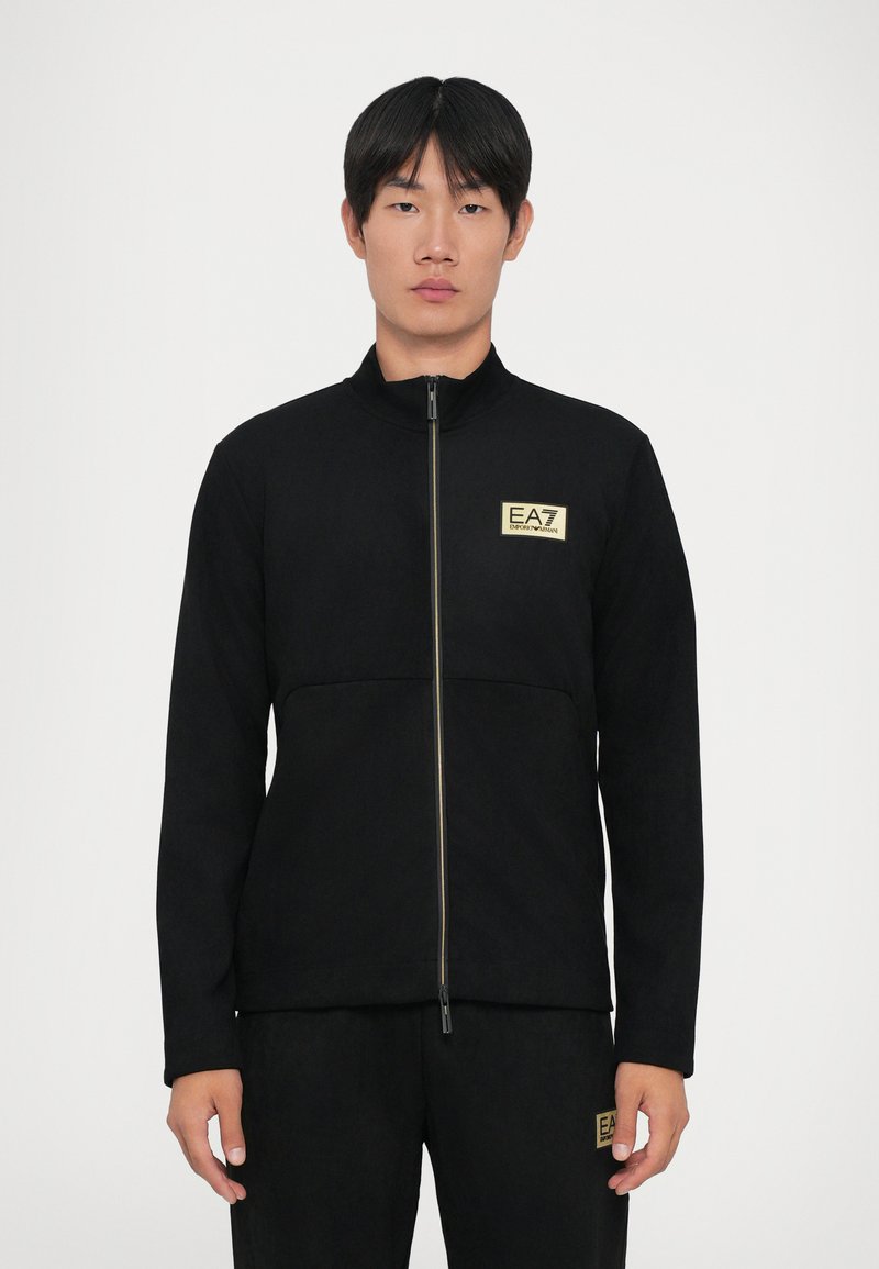 Uomo che indossa una giacca nera con zip e pantaloni con toppe del logo EA7 Emporio Armani sul petto e sulla coscia, in piedi davanti a uno sfondo semplice.