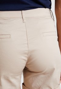 Shorts en coton beige avec une texture lisse, dotés de poches arrière et de coutures cousues. La taille est simple, conçue pour un ajustement décontracté.