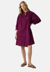 Robe violette à boutons avec une coupe décontractée, manches bouffantes trois-quarts et ourlet arrivant au genou ; accompagnée de sandales à imprimé léopard.