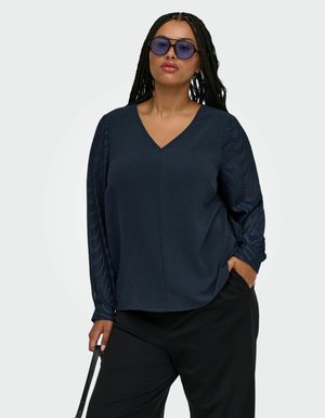 Donna con lunghi capelli intrecciati che indossa occhiali da sole blu, una blusa blu scuro con maniche trasparenti decorate, e pantaloni neri, che tiene una tracolla in una mano.