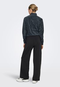 Γυναίκα με τα μαλλιά της σε κότσο, που φοράει ένα σκούρο μπλε φλις μπουφάν με λεοπάρ σχέδιο, μαύρο παντελόνι με φαρδιά πόδια και άσπρα αθλητικά παπούτσια, όρθια, με την πλάτη προς τα εμάς.