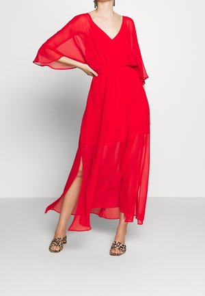 Kobieta w zwiewnej czerwonej sukni z dekoltem w kształcie litery V, przezroczystymi rękawami i rozcięciami po bokach, w połączeniu z sandałami na obcasie z odkrytymi palcami w panterkę, stojąca na białym tle.