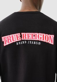 Schwarzer Pullover mit dem großen roten und weißen Schriftzug "TRUE RELIGION" auf dem Rücken und dem kleineren weißen Schriftzug "BRAND JEANS" darunter.