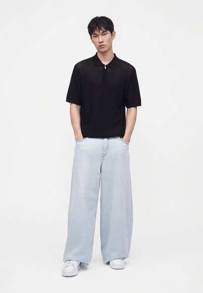 Jeune homme portant un t-shirt noir à manches courtes avec un col zippé, un jean large bleu clair et des baskets blanches, mains dans les poches.