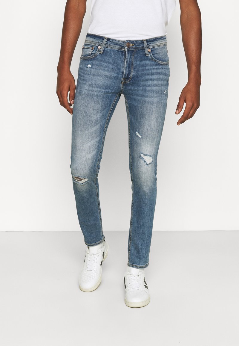 Ublekta blå denimjeans med smal passform, med slitna detaljer och synliga slitningar, i kombination med vita sneakers.
