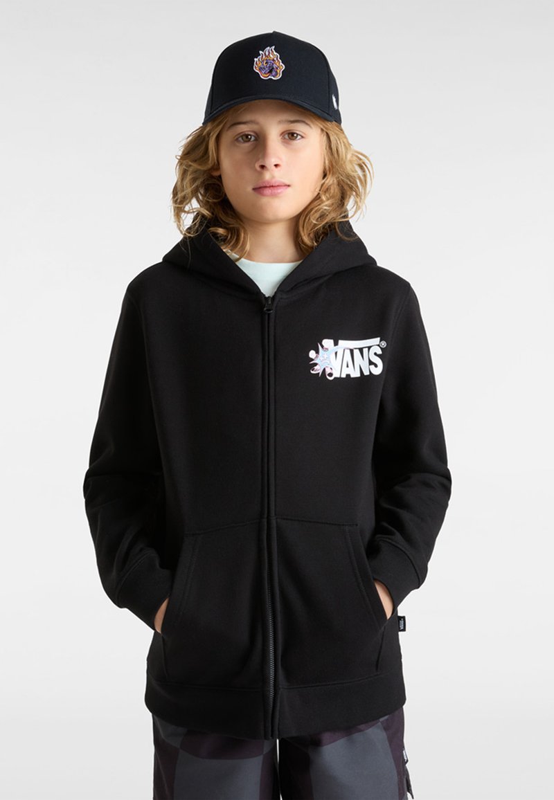 Haut à capuche noir zippé avec poches avant et logo Vans blanc. Porté sur une chemise bleu clair, associé à un short à motifs sombres et une casquette.