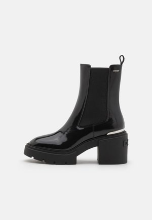 Bottine en cuir verni noir à talon bloc épais, avec panneau latéral élastique, languette, et logo DKNY argenté sur le côté supérieur.