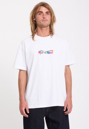 Jeune homme aux cheveux blonds mi-longs porte un t-shirt blanc oversize avec un motif abstrait coloré sur la poitrine et un pantalon sombre.