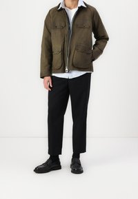 Mann, der eine olivgrüne Reißverschlussjacke mit vier vorderen Taschen, schwarze 3/4-Hosen, schwarze Socken und schwarze Slipper trägt und vor einem weißen Hintergrund steht.