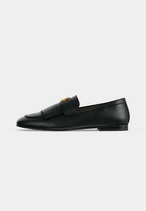 Mocassin en cuir noir avec un rabat avant à franges et une petite boucle dorée sur un fond gris clair uni.