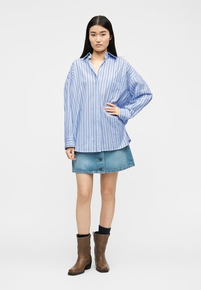 Γυναίκα όρθια με το χέρι στη μέση, φοράει ένα φαρδύ πουκάμισο με μπλε ριγέ κουμπιά, φούστα από τζιν, μαύρες κάλτσες και καφέ μποτάκια στον αστράγαλο.