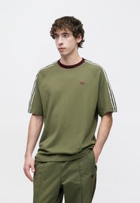 Φούτερ σε ελιάς πράσινο με μαρουν πλεκτό γιακά και τρεις λευκές ρίγες στους ώμους. Διαθέτει μικρό βουργουνδί λογότυπο στο στήθος.