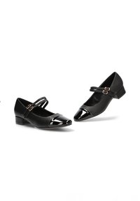 Zapatos negros con acabado mate y punteras de charol brillante. Presentan correas en el tobillo con hebillas en tono dorado y tacones bajos en bloque.