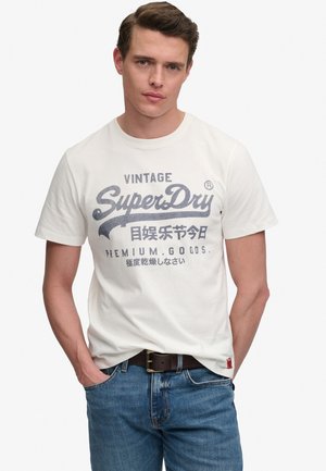 Jeune homme aux cheveux courts et bruns portant un t-shirt blanc avec le logo vintage SuperDry, un jean bleu et une ceinture marron, les mains dans les poches.