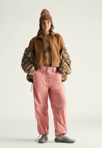 Braune cropped Zip-Jacke, rosa Hose, braune Strickmütze. Strukturiertes Oberteil, weite Ärmel, reflektierende Akzente an der Hose, sportliche Schuhe mit grauen Details.