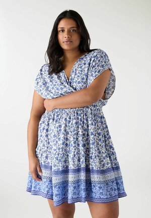 Donna con lunghi capelli scuri che indossa un vestito corto a fiori blu e bianco, in piedi con un braccio posato sulla vita.