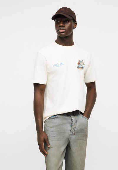 Άνδρας με λευκό μπλουζάκι με σχέδιο, ανοιχτόχρωμο τζιν και σκούρο καφέ καπέλο, στέκεται με το ένα χέρι στην τσέπη μπροστά σε απλό φόντο.