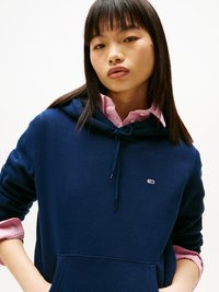 Mørkeblå hettegenser med frontlomme, snorhette og en liten logo. Kombinert med en lys rosa skjorte med krage under.