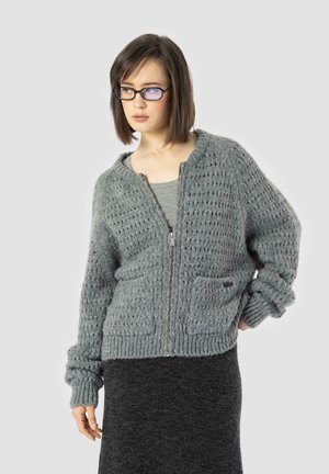 Grauer gestrickter Cardigan mit Reißverschluss, gerippter Struktur, lockerer Passform und zwei Vorderentaschen. Kombiniert mit einem dunkelgrauen Rock.