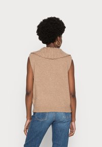 Maglione in maglia beige senza maniche con colletto alto a coste e una vestibilità rilassata, abbinato a jeans in denim blu. Tessuto morbido e design casual.