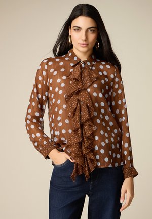 Vrouw met lang donker haar draagt een bruine blouse met witte stippen en een gerafelde voorkant, gecombineerd met donkerblauwe jeans, staand voor een beige achtergrond.