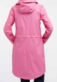 Henkilö, joka käyttää pitkää, huppupäistä vaaleanpunaista sadetakki, näkyy selkäpuolelta, kädet lanteilla yksinkertaista taustaa vasten.