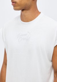 Άνδρας που φοράει λευκό αμάνικο μπλουζάκι Puma Crew '48, εμφανιζόμενος από τον λαιμό έως το πάνω μέρος του στήθους και τους ώμους, σε απλό φόντο.