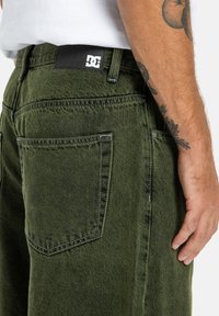 Zielone dżinsy o klasycznym kroju, z widocznym szwem, czarną metką DC na pasku oraz dwiema tylnymi kieszeniami.