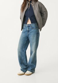 Casual outfit met een blauwe ritssluiting crop top, grijze gewatteerde jas, losse blauwe jeans en witte sneakers met beige accenten, in een staande pose.
