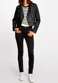 Zwarte leren motorjack met ritsen. Gestreept zwart-wit langarmshirt eronder. Slim-fit zwarte broek en witte sneakers.