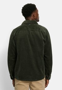 Dark green corduroy shirt with a ribbed texture, button-down collar, and long sleeves, featuring a subtle seam detail across the back.