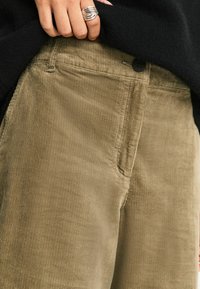 Khakifarbene Cordhose mit Rippenstruktur, mit einer vorderen Tasche und einem schwarzen Knopfverschluss. Das Oberteil ist in den Bund gesteckt.