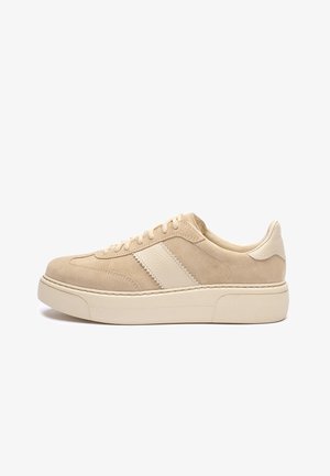 Beżowy zamszowy trampki z teksturowanym białym skórzanym paskiem, okrągłym noskiem i grubą kremową gumową podeszwą. Sznurowadła i przeszycia są w kolorze beżowym, dopasowane.