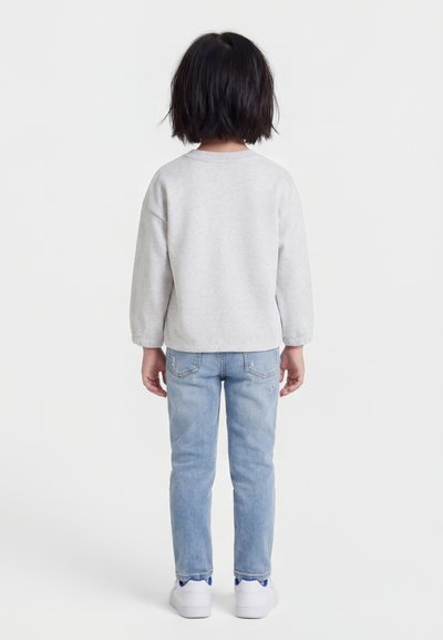 Otrok s kratkimi črnimi lasmi stoji obrnjena stran, oblečen v svetlo sivo trenirko, svetlo modre kavbojke in bele superge na belem ozadju.