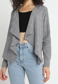 Černobílý kostkovaný otevřený kabát s drapovaným designem, dlouhými rukávy a měkkou látkou, doplněný černým crop topem a džínami s vysokým pasem.