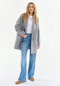 Žena oblečená v sivej oversized blejzri so vzorom rybej kosti, bielom tričku, modrých širokých džínsach, bronzových topánkach a náhrdelníku z korálok, stojaca na bielom pozadí.