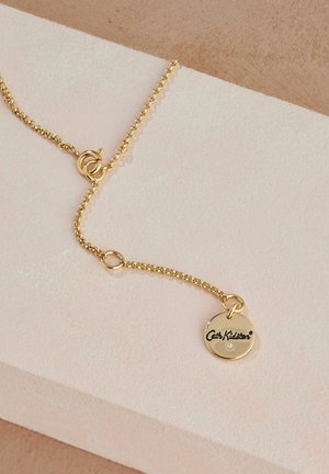 Gold chain necklace with a round pendant featuring the engraved text "Cath Kidston" and a small gem accent. Smooth texture and shiny finish.