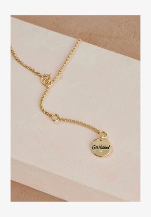 Zelta ķēde ar apaļu piekariņu, uz kura ir iegravēts teksts "Cath Kidston" un neliela dārgakmens akcenta. Gluda tekstūra un spīdīga apdare.