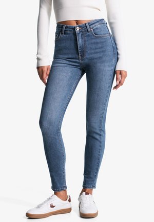 Jeans en denim bleu skinny à taille haute, avec une finition délavée, un design à cinq poches et des détails de couture subtils, associés à des baskets blanches.