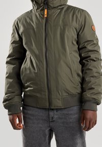 Olivově zelená prošívaná bunda s předním zipem, kapucí a žebrovanými manžetami. Má výrazný oranžový táhlo zipu a logo na rukávu.
