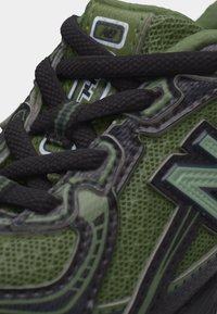 Πράσινο αθλητικό παπούτσι από διχτυωτό ύφασμα με μαύρα κορδόνια, ανάγλυφη σόλα και στυλιζαρισμένο λογότυπο στο πλευρό κοντά στις ενισχυμένες σειρές οπών.