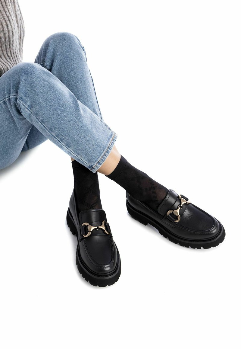 Zwarte leren loafers met een gouden kettingaccent, dikke zool, gedragen met zwarte gestreepte sokken en blauwe opgerolde denim jeans.